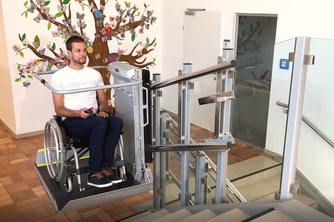 Stopniščna dvižna ploščad prepelje osebo na invalidskem vozičku čez stopnišče. 