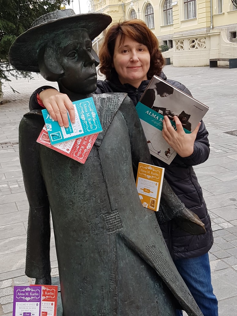 Jerneja Jezernik ob Almi Karlin (kip v Celju je delo Vasilija Ćetkovića) z njunimi knjigami. Fotografija je bila posneta natanko ob 100. obletnici odhoda Alme M. Karlin iz Celja na njeno pot okrog sveta, 24. novembra 2019. 