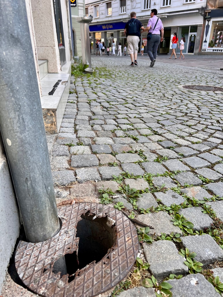 Poškodovani pokrov jaška na Grajskem trgu 7, tik ob stavbi in kovinskem drogu. 