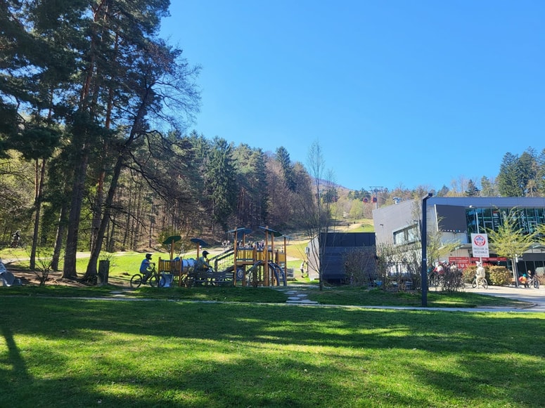 Kolesarji med vožnjo skozi območje z otroškimi igrali pri spodnji postaji vzpenjače. 